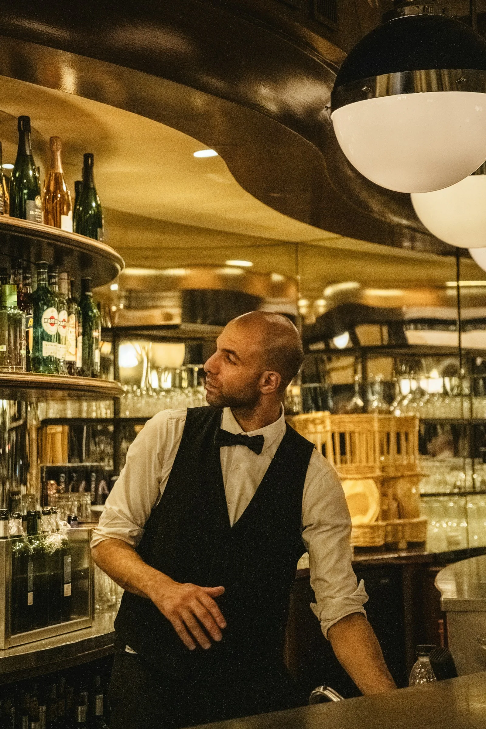A bar manager in a black vest and bow tie standing behind a classic restaurant bar with rows of bottles and warm pendant lighting — the kind of composed leadership that defines a strong manager